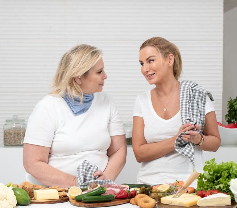 Two women in a kitchen preparing fresh vegetables and cheese with olive oil and spices.