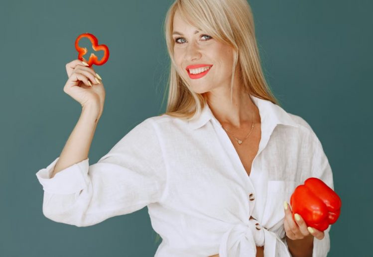 Blonde woman in white shirt smiling while holding a fresh red bell pepper slice.