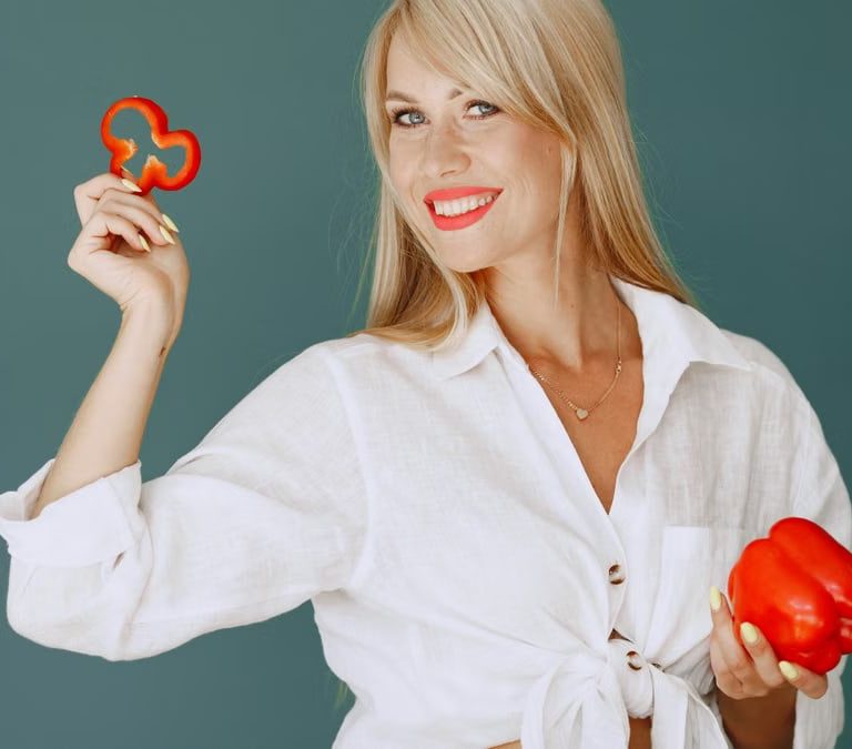 Blonde woman in white shirt smiling while holding a fresh red bell pepper slice.